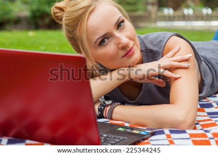 Sensual blonde woman lying in park on blanket. She is using red laptop pc. Outdoor photo. She looks relaxed.