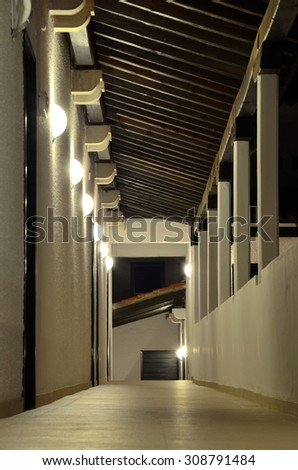 Semi-open corridor under a wooden roof, with a succession of the wall lamps on the left side, and columns on the right side. Corridors and passageways under artificial lighting (reprocessed image)