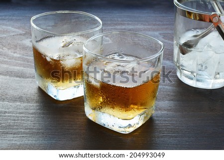 Two glasses of whiskey over ice with ice bucket on wooden table.