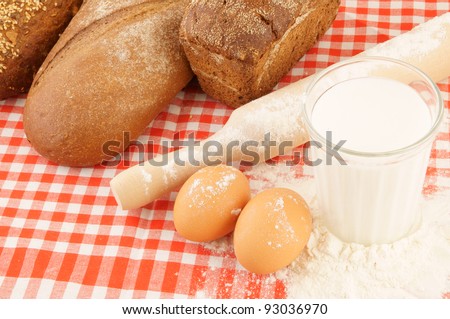Flour, milk, eggs, bread and rolling pin on red checked tablecloth