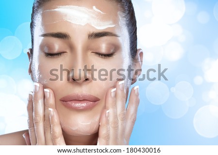 Beautiful healthy young woman with eyes closed while applying moisturizer to her clean face using gentle soft touches, close-up portrait on blue and white background