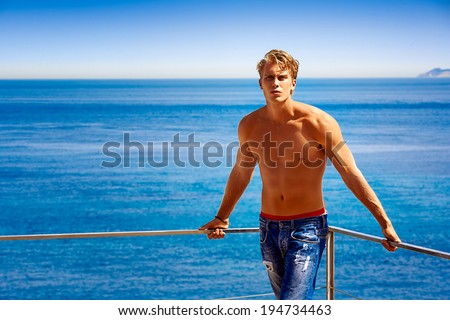 Dream Vacation. Handsome blonde young man in jeans and shirtless sunbathe at the balcony with sea background