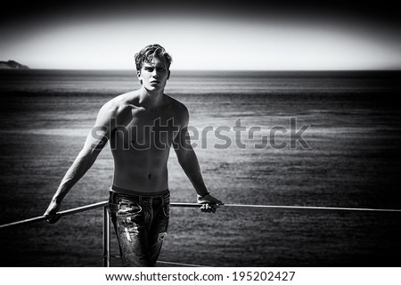Dream Vacation in Black and White. Handsome blonde young man in jeans and shirtless sunbathe at the balcony with sea background