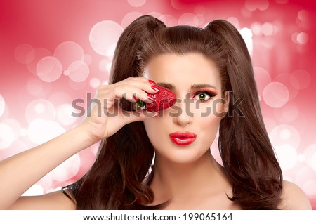 Happy and young woman holding a strawberry in eye with funny expression. Professional make-up and nails in red.