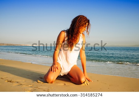Pretty Young Woman Kneeling Down on the Beach Against Peaceful Ocean under a Blue Sky in the Sunset.