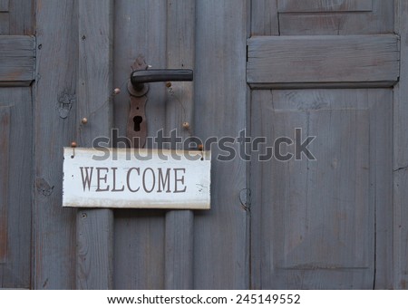 Welcome sign hanging below the handle on an old grey wooden door greeting guests as they enter