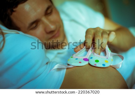Image of happy future dad holding baby shoes on the belly of his caucasian pregnant wife lying on bed at home.