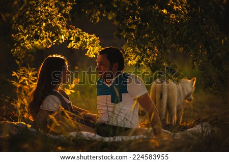 Happy smiling couple relaxing on green grass in the evening. Family is resting in the garden or park into the sunset. Young man looks at a beautiful girl.