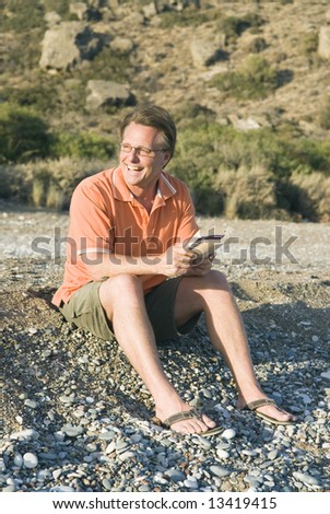 A happy smiling 44 year old man wearing spectacles is reading book on beach.