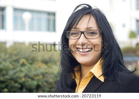 A beautiful indian business woman wearing spectacles gives a stunning smile as she stands in front of office buildings