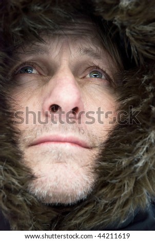 A color portrait photo of a handsome mature man with stubble wearing a furry hooded winter coat.