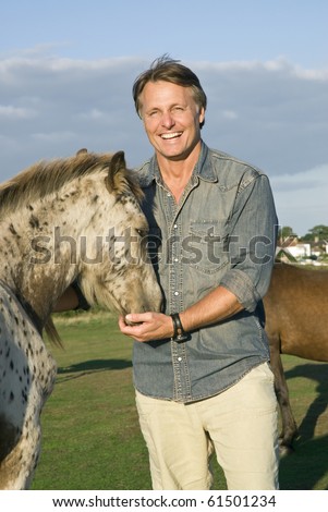 A color portrait photo of a happy smiling forties man man petting his young appaloosa horse.