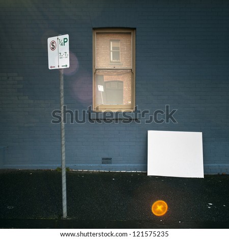 Parking sign on pavement with blank white board, blue wall and window. Surreal perspective and strong lens flare effect.