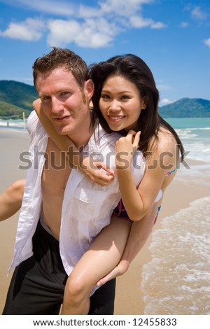 caucasian man & asian woman having fun carrying piggyback at the beach