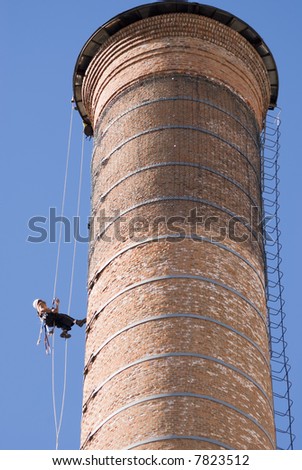 Cropped shot of an unrecognizable worker hanging from a high industrial brick chimney. Blue sky in the background.