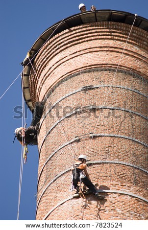 Cropped shot of two unrecognisable workers hanging from a high industrial brick chimney. Blue sky in the background.
