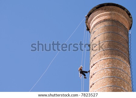Cropped shot of an unrecognizable worker hanging from a high industrial brick chimney. Blue sky in the background, with huge copy space.
