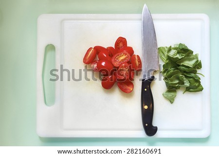 High angle shot of kitchen knife resting on white chopping board with cherry tomatoes in half and fresh basil leaves.