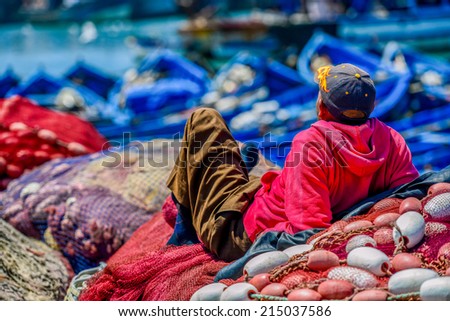 A old man with straw hat sitting in th middle of red fishing nets spread on the ground and sewing nets