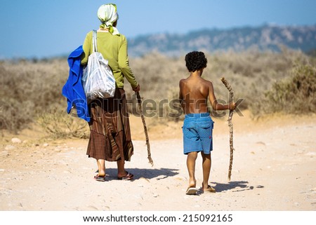 An old woman and a young boy walking along the dusty road in the countryside towards the hill