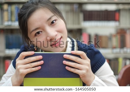 Portrait of clever girl student resting her chin on a text book in front of a library bookshelf