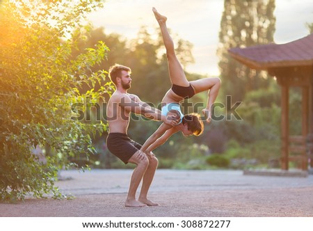 Acro yoga, two sporty people practice yoga in pair, couple doing stretching exercise in the garden with evening sunset on background