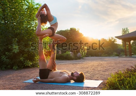 Acro yoga, two sporty people practice yoga in pair, couple doing stretching exercise in the garden with evening sunset on background