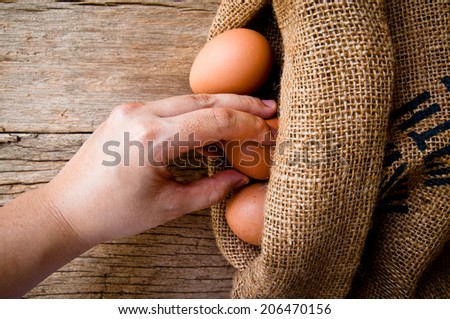 Hand Holding (Select,Pick,Choose) Fresh Egg With Burlap Sack Harvest on Wooden Table Background, Food Rustic Still Life Style. Concept and Idea for Homemade Food Art Decoration.