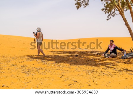 Couple of tourists resting under a tree on sand dunes in Merzouga, Morocco