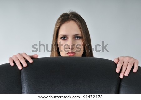 Studio portrait of young woman hidden behind big black armchair - face with hands visible