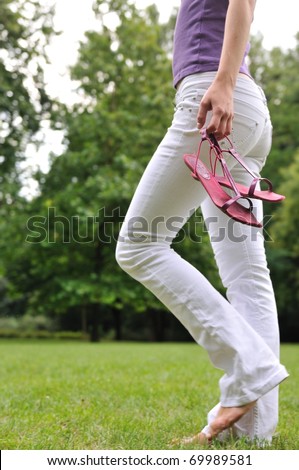 Woman (torso) walking barefoot on grass in park and holding her shoes in hand