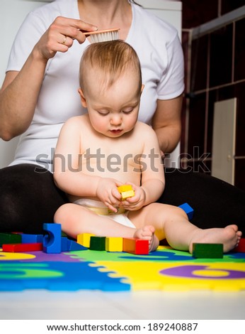 Mother brushing hair of her cute baby while she is playing with toys in bathroom