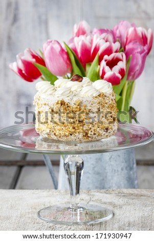 Round nut cake on cake stand. Beautiful bouquet of pink and red tulips in the background.