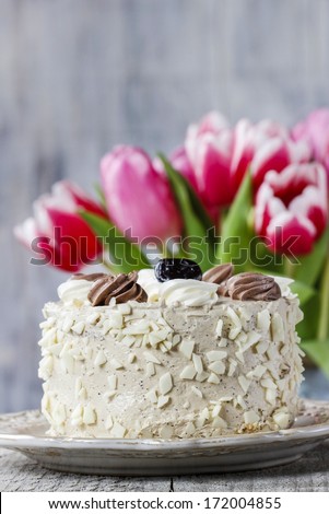 Round cappuccino cake on cake stand. Beautiful bouquet of pink and red tulips in the background.