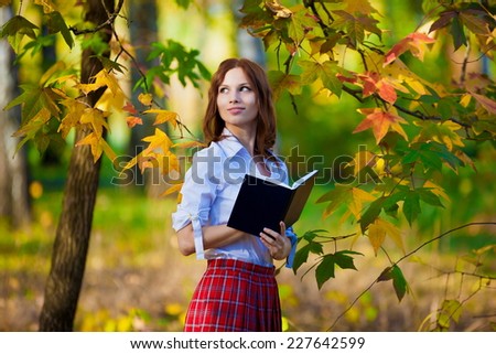 beautiful  smiling  schoolgirl in red skirt and white blouse posing in autumn nature has book