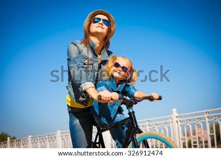Beautiful mother and daughter cyclist, has happy fun cheerful smiling face, yellow t-shirt, blue jeans and hat, sunglasses, sport body. Motion on great bicycle in urban city. Portrait nature.