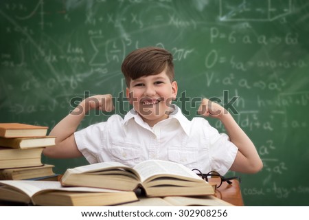 Smart and happy  boy sitting at desk  front of blackboard  expressing the power of knowledge. Educational concept. Copy space.