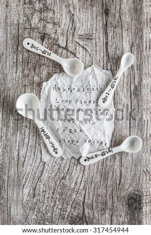 Measuring tablespoons and teaspoons on a wooden board along with a torn piece of paper containing the ingredients for a recipe.