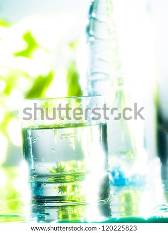 A transparent glass of fresh water with a plastic bottle and green plants in the background. The transparency is enhanced by the backlight and the blurred effect in the background.
