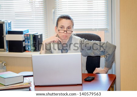 A fifty years old and emotional woman wearing glasses and working in office with a laptop is very skeptical with the modern technology. She looks at the monitor with confusion.