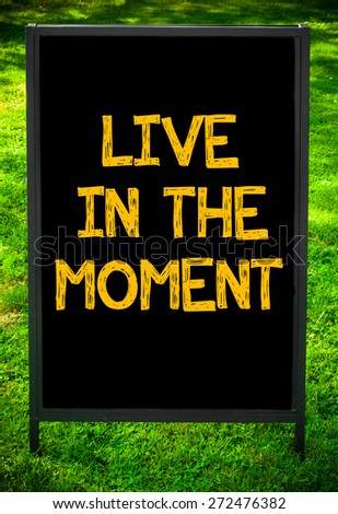 LIVE IN THE MOMENT  message on sidewalk blackboard sign against green grass background. Copy Space available. Concept image
