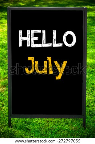 HELLO JULY  message on sidewalk blackboard sign against green grass background. Copy Space available. Concept image