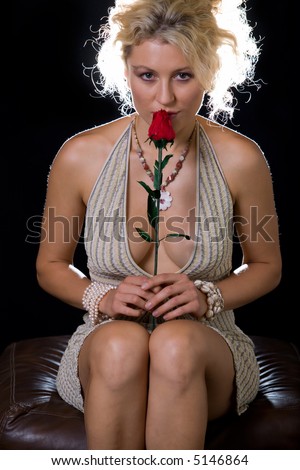 Full body of an attractive woman with curly blond hair in a sexy low cut dress sitting over black holding a red rose