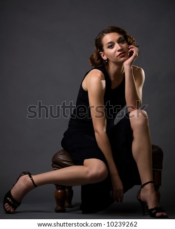 Full body of young pretty brunette woman modeling long black dress while sitting on a stool looking bored on grey background