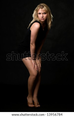 Full body of an attractive young blond woman wearing short black dress standing with hands on knees over black with hair back lit