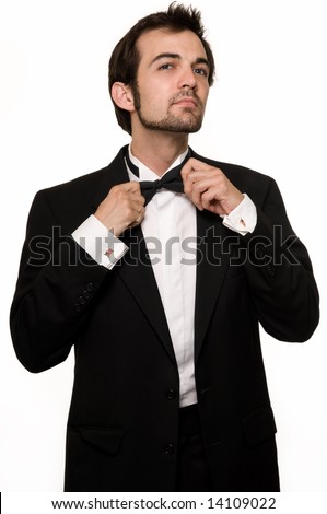 Attractive young brunette man with a beard wearing a black tuxedo fixing bow tie standing on white