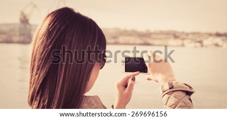 Fashion  woman taking photo with cellphone on the coast on spring. Happy girl on vacation taking picture on sea background.