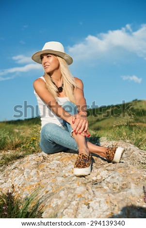 portrait of the attractive, slender, beautiful young Caucasian  blonde girl in a stinny hat.Smiling girl enjoys fine warm sunner weather highly in mountains against the sea