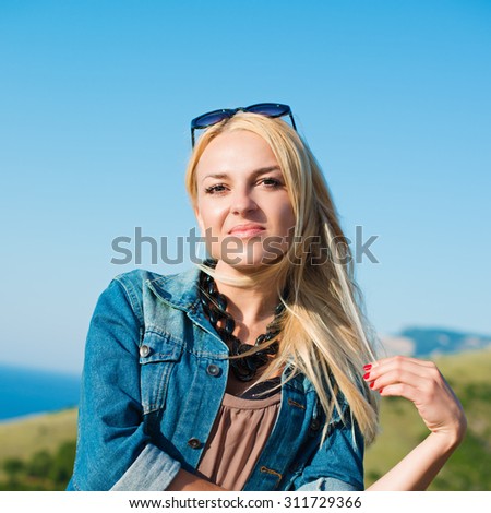 portrait of the attractive, slender, beautiful young Caucasian  blonde girl in a jeans jacket. Smiling girl enjoys fine warm sunner weather highly in mountains against the sea