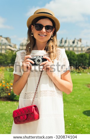 young attractive woman in hat, white dress, red bag and retro camera poses against Paris. Fashion and city style.